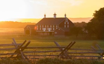A scenic view of a barn at sunset, surrounded by fields and a wooden fence.