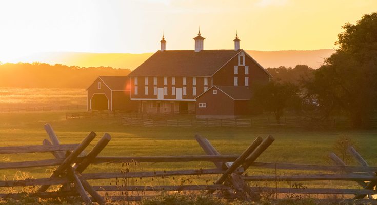 A scenic view of a barn at sunset, surrounded by fields and a wooden fence.
